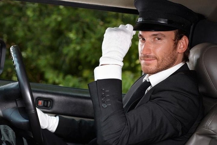 A professional chauffeur in a black suit and white gloves inside a luxury car, offering Car Rentals With Drivers service.